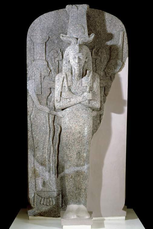 A large red granite lid of sarcophagus. There are three images of people carved into the granite, standing up. The central figure is male and protrudes from the lid, facing the viewer. His arms are crossed across his chest and he wears a beard, headpiece, and crown of feathers and horns. The two women either side of him face him and have their hands on his waist. The lid is broken on the right, and the side and the feet have been filled in in white plaster.