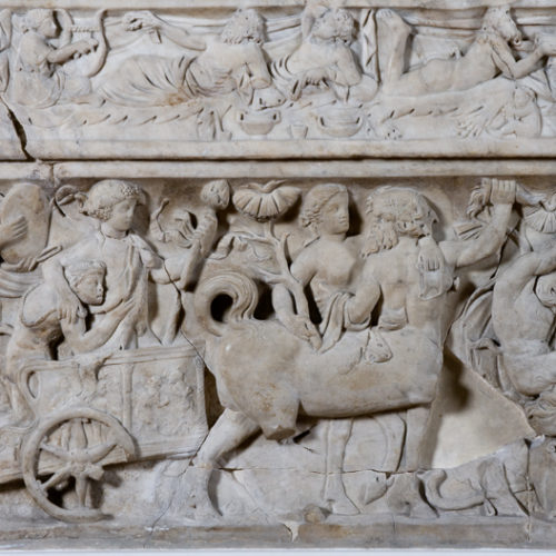A photograph of a section of a highly sculpted marble coffin, showing a person riding a chariot as well as two centaurs, as well as people relaxing and eating all across the top.