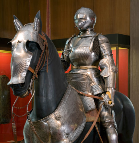 A mannequin dressed in a medieval suit of armour sits on a lifesize horse model, which is also dressd in armour