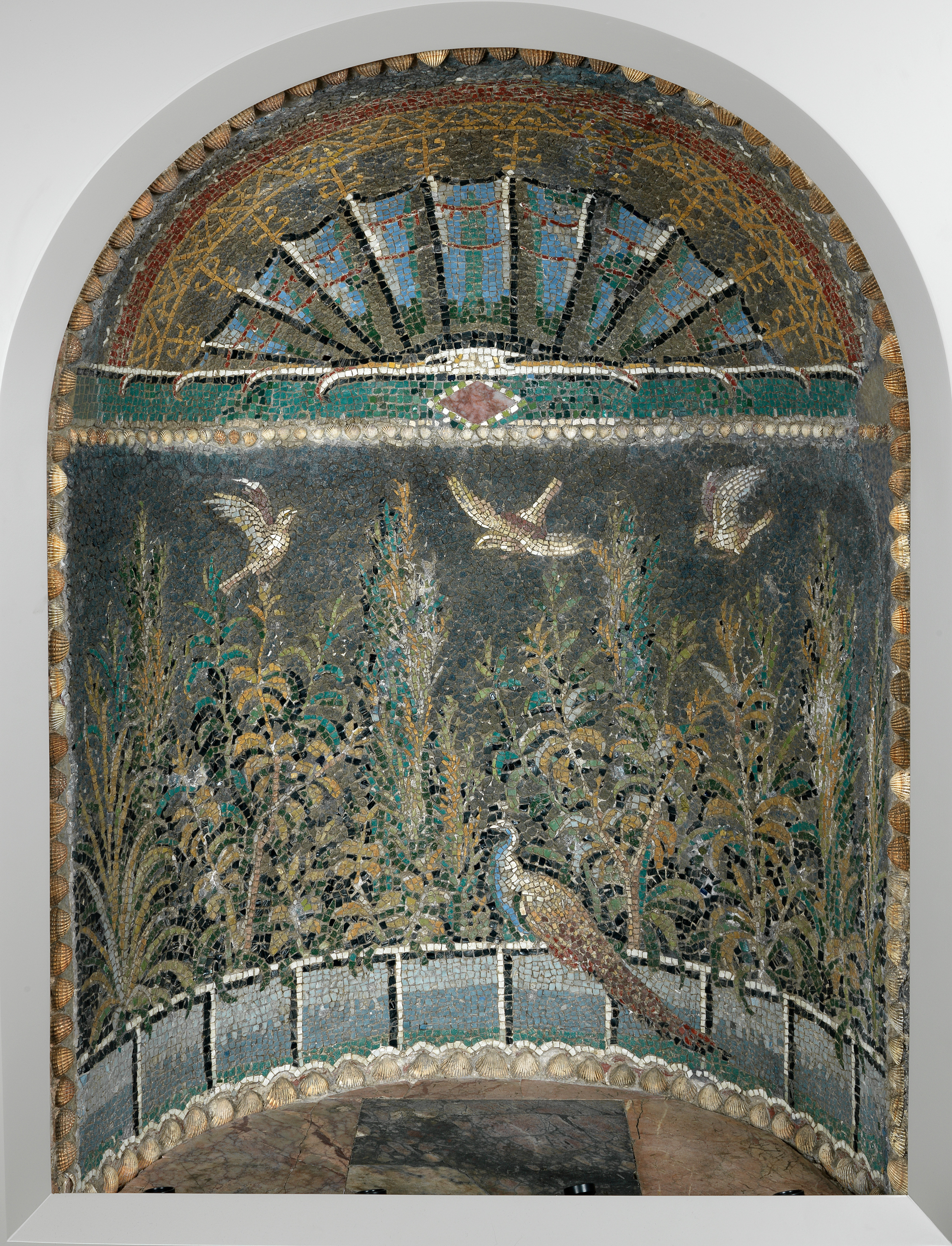 Photograph of a niche with an ancient Roman mosaic in it. The mosaic shows plants, three birds flying at the top and a peacock perched on the edge at the bottom.