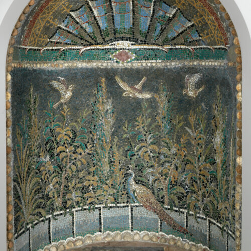 Photograph of a Roman mosaic laid in a niche. The colours are blues and greens and an optical illusion is created of a window that looks out onto a scene of plants, flying birds, and one peacock perching on the ledge.