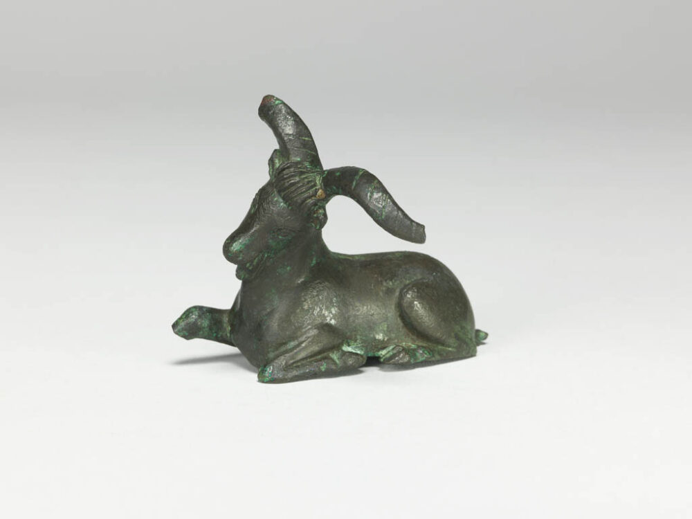 A photo of a small bronze goat statuette against a white backdrop. The goat is lying down, with their head and horns tilted to its left. One of it's front legs is lifted up and a little bit has broken off the end.