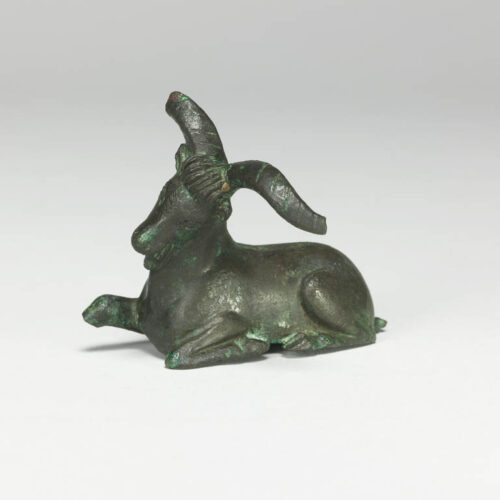 A photo of a small bronze goat statuette against a white backdrop. The goat is lying down, with their head and horns tilted to its left. One of it's front legs is lifted up and a little bit has broken off the end.
