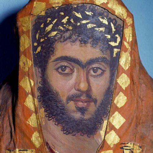 A close up photograph of a painted portrait of an Egyptian man's face, with thick curly beard and moustache, and thick strong eyebrows. He is also wearing a golden painted wreath in his hair, and the portrait is surrounded by orange-red mummy wrappings.