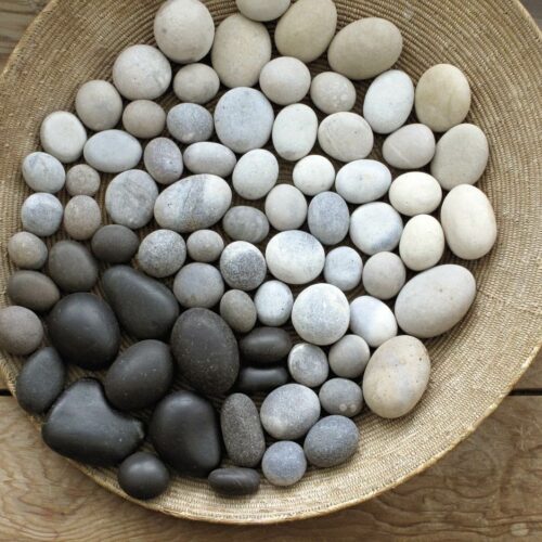 A photograph of a basket filled with smooth round pebbles. The pebbles are arranged: from bottom left they are larger and dark grey in colour. They change colour across the basket, from dark grey, to lighter grey, to white. Larger pebbles are around the edge.