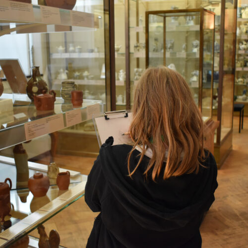 A girl draws on a clipboard whilst looking at some ceramics in a glass cabinet.