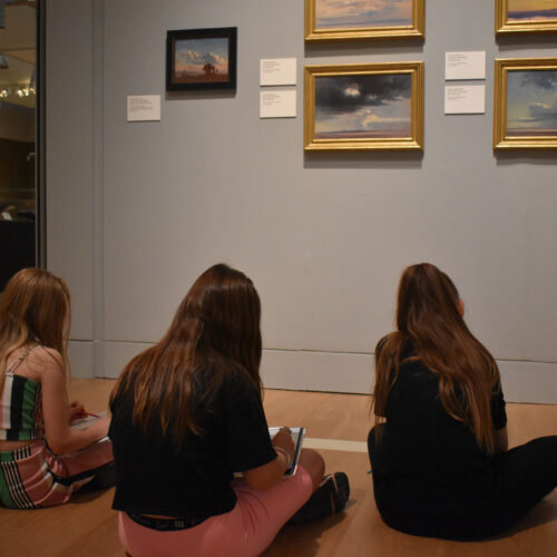 Three children sketching whilst looking at paintings in a museum gallery.