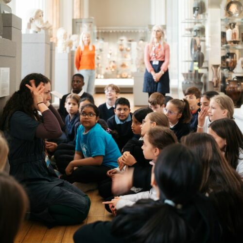 group of primary school children seated on floor of ancient Greek gallery, in front of adult female who is animatedly talking