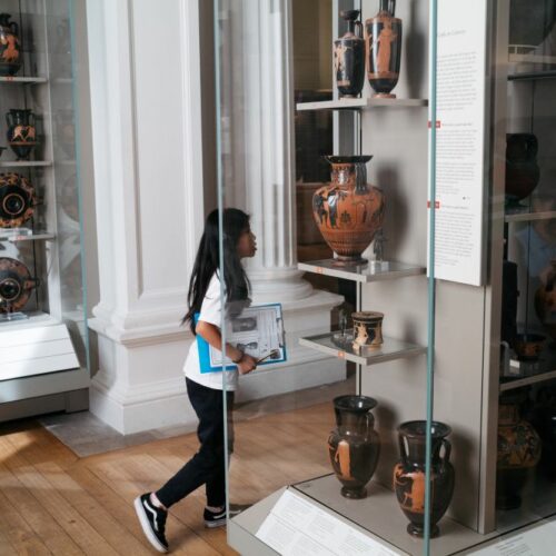 young primary age girl walking with a clipboard in the ancient Greek gallery, cases of Greek vases in foreground and background